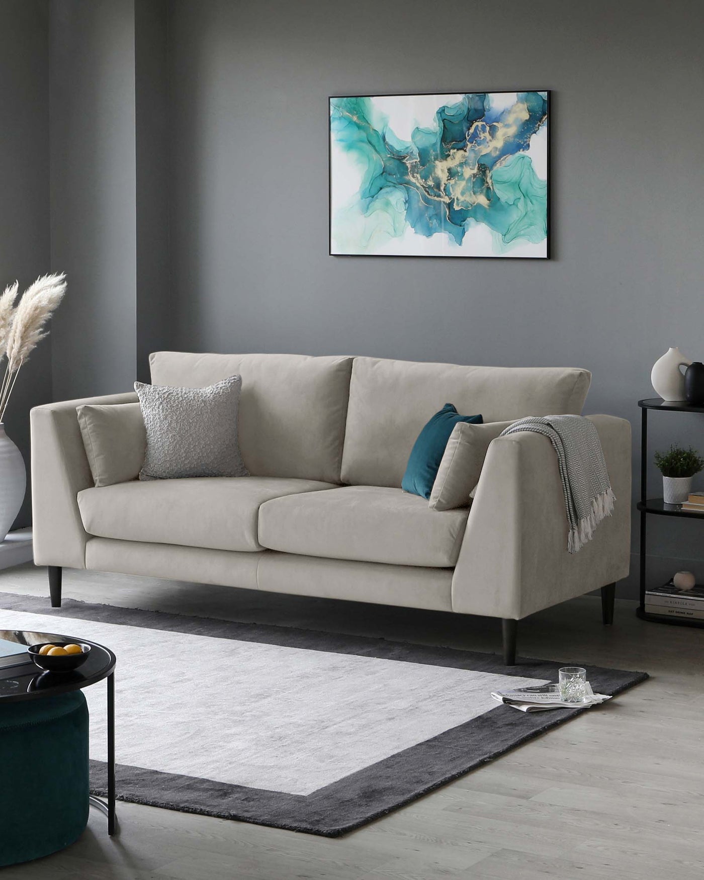 A modern three-seater sofa in a neutral colour with minimalist arm design and clean lines, accompanied by a teal ottoman, a two-tier round coffee table with a matte black finish, and a two-tone grey area rug. Decor includes assorted decorative cushions, a light grey throw blanket, and a small side table with a monochromatic vase. The scene is set against a grey backdrop, highlighting the furniture's contemporary aesthetic.