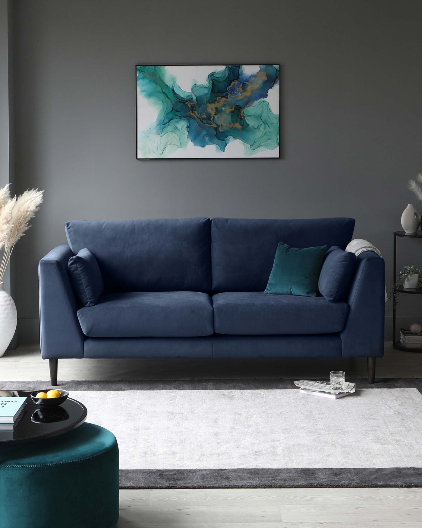 Elegant deep blue fabric three-seater sofa with plush cushions and flared armrests, accompanied by a round teal velvet ottoman, resting on a modern grey and white area rug.