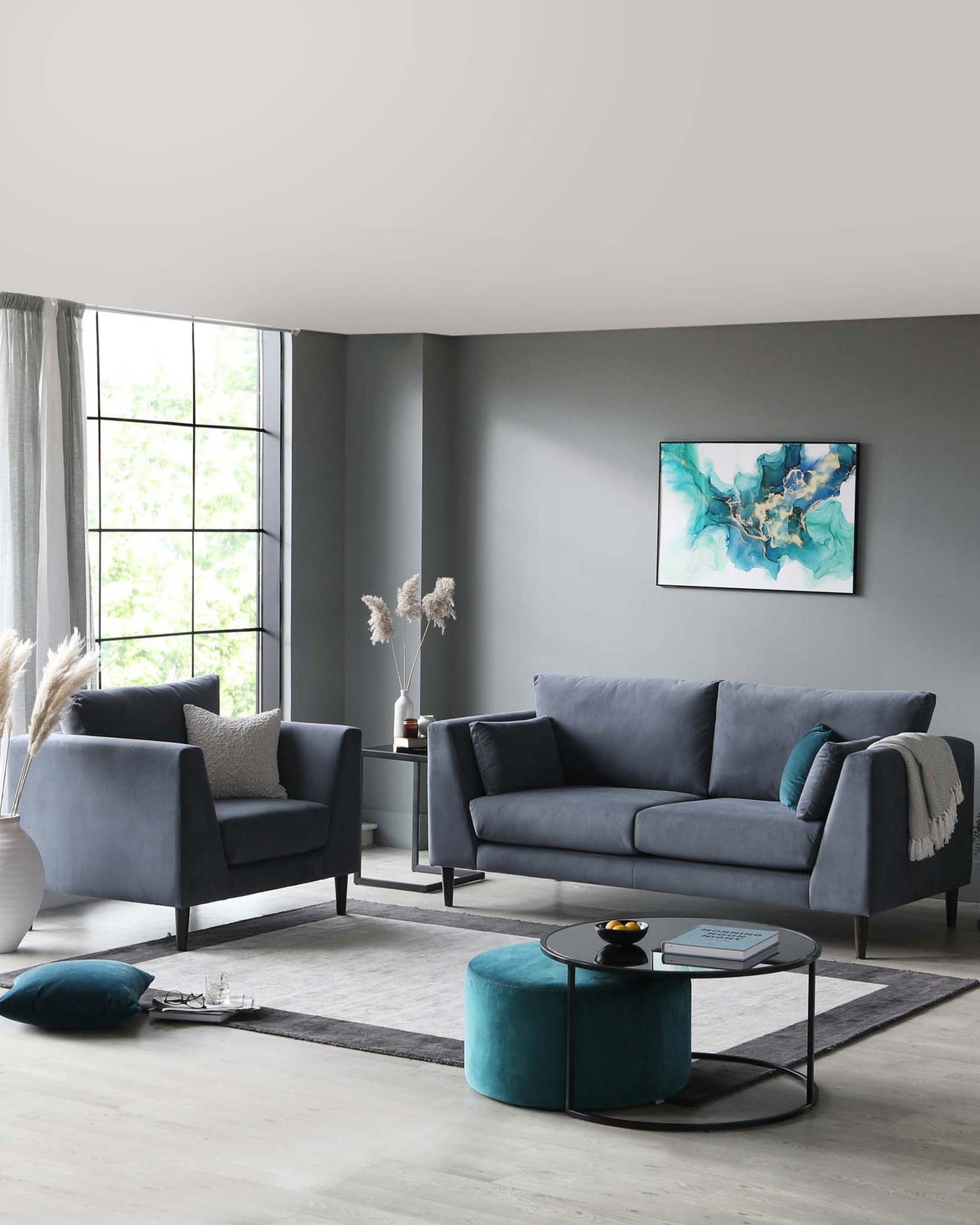 Modern living room set with a dark grey upholstered sofa and two matching armchairs. A round, black metal-framed coffee table with a glass top centres the arrangement, complemented by a teal tufted fabric ottoman. The furniture rests on a light grey area rug over a pale wood floor.