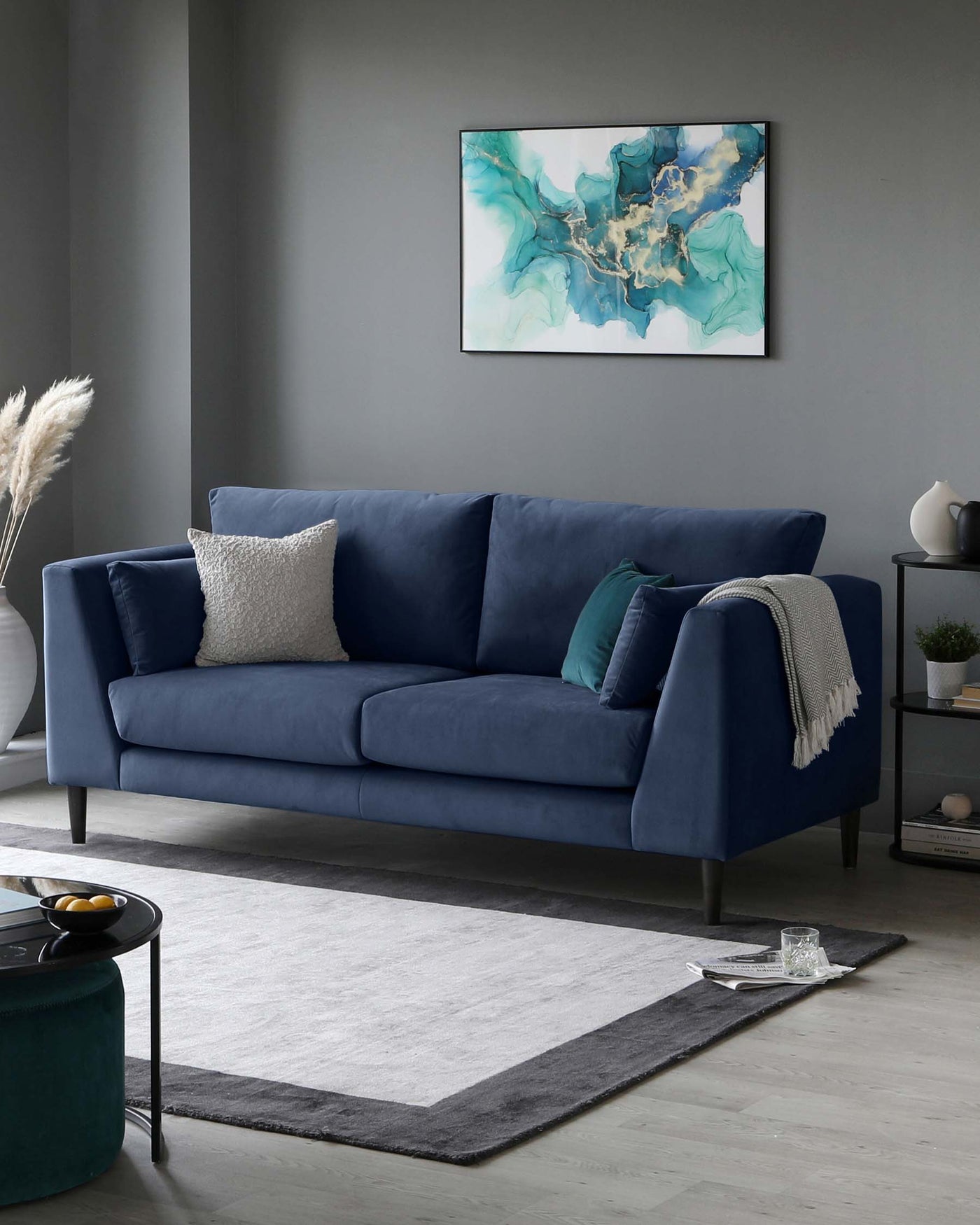 Elegant modern living room with a plush velvet three-seater sofa in a deep navy blue, featuring clean lines, tapered legs, and including assorted throw pillows in matching and contrasting colours. A dark green round velvet ottoman sits in front of the sofa, while to the right, a minimalist grey sideboard displays decorative items and a sleek white vase. The ensemble is anchored by a large two-tone geometric area rug in grey and white, complementing the room's contemporary aesthetic.