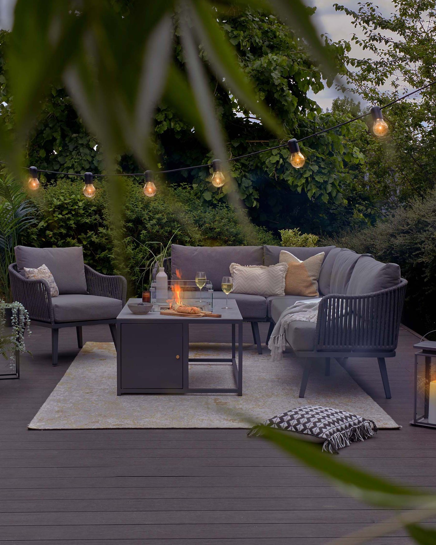 Outdoor furniture set on a wooden deck, including a dark grey sectional sofa with plush cushions, complemented by matching armchairs. A rectangular metal fire pit table sits at the centre, flanked by glassware. Soft lighting from overhead string bulbs adds a cosy ambiance. An off-white patterned area rug lies beneath the set, and accessories like a throw blanket and decorative pillows enhance the overall comfort. Transparent lanterns contribute to the evening's tranquil mood.