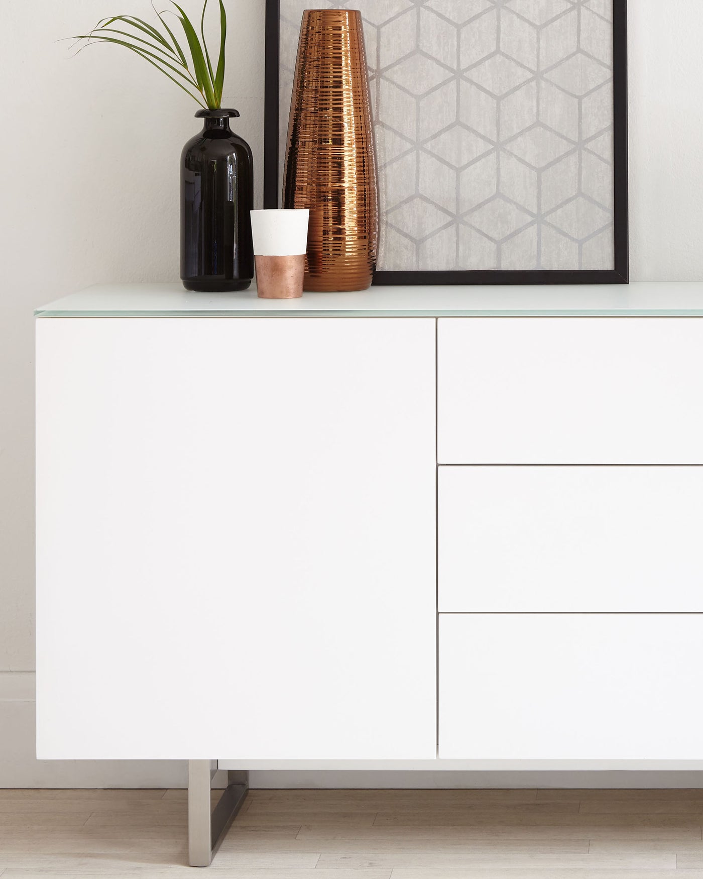 Modern minimalist sideboard with a matte white finish and sleek metal legs. The sideboard features one large cabinet door and three drawers on the right side, offering a blend of concealed and organized storage. It is topped with a frosted glass surface, complemented by decorative items including a tall copper vase, a small plant in a black vase, and a framed geometric artwork resting against the wall. The design showcases clean lines and a contemporary aesthetic ideal for modern living spaces.