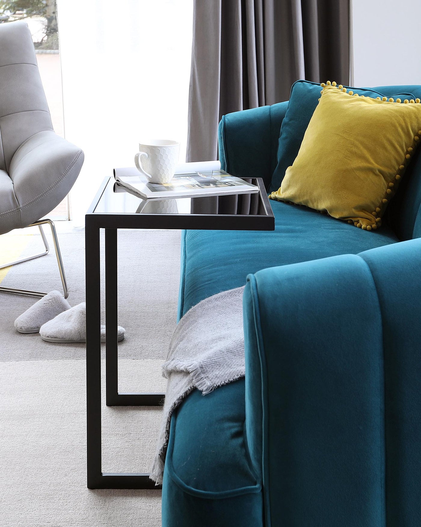 A modern living room setting featuring a plush teal sofa adorned with a vibrant yellow decorative pillow and a light grey throw blanket draped casually on one side. In front, a sleek, minimalist black metal C-table with a rectangular top holds a white cup and a magazine, offering both style and functionality. A grey upholstered accent chair with a curved back and angled metal legs provides additional seating, enhancing the contemporary aesthetic of the space.