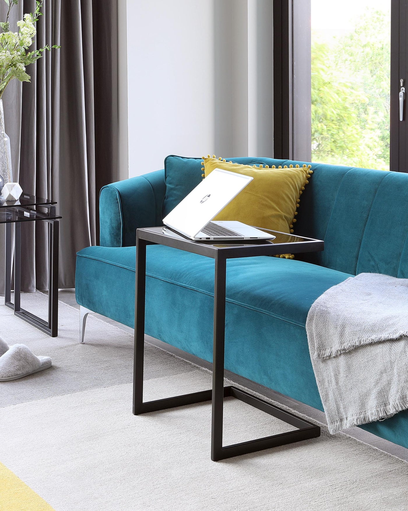 Contemporary teal fabric sofa with tufted backrest and brass-studded detailing, paired with a sleek black metal frame side table featuring a glass top.