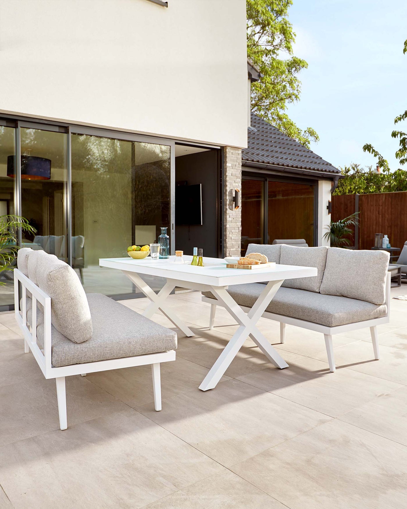 Modern outdoor furniture set featuring a white rectangular dining table with a minimalist design and criss-cross legs, paired with two armless, light grey upholstered dining chairs with white frames. The set is arranged on a beige tiled patio.