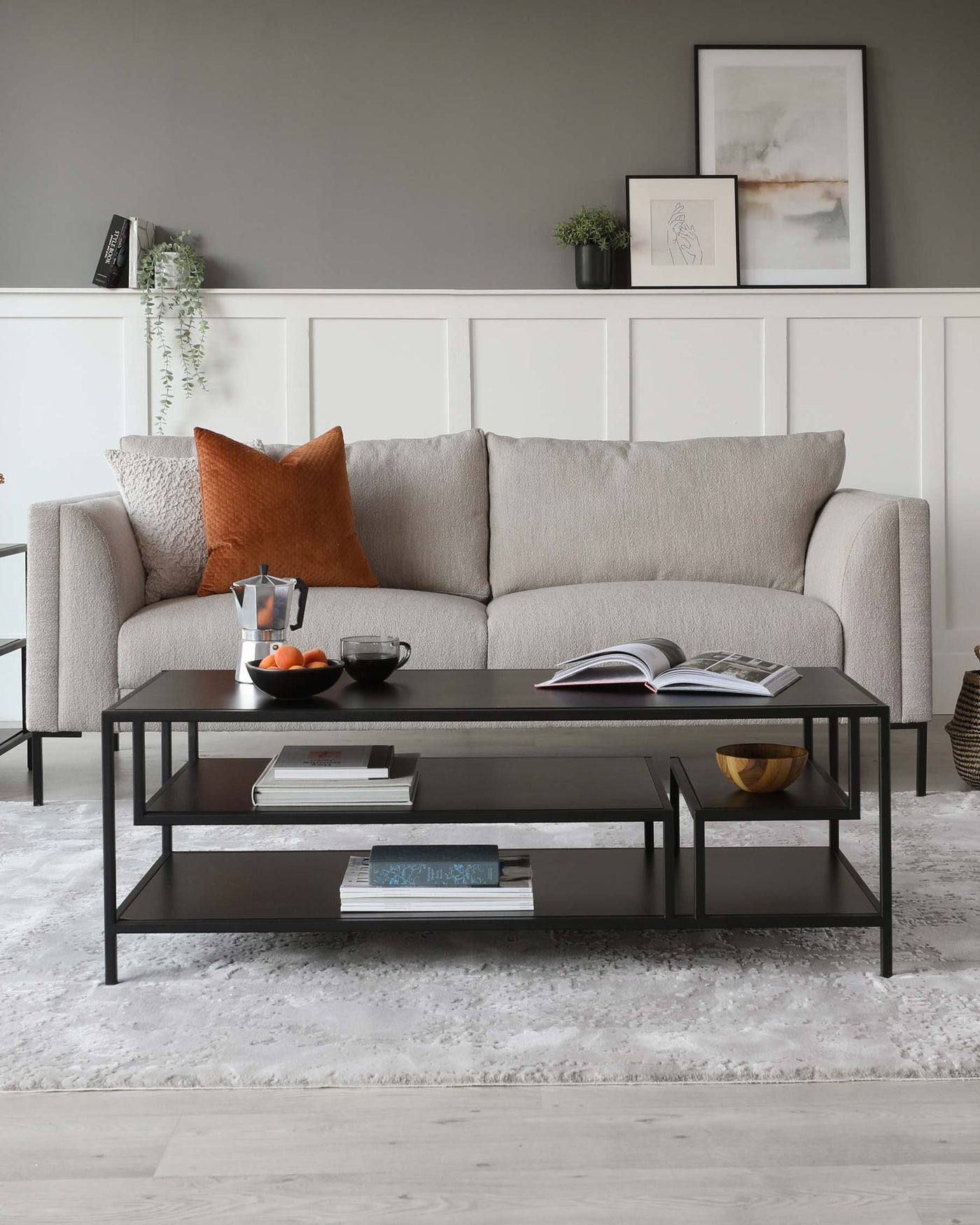 A contemporary living room featuring a neutral-toned upholstered three-seater sofa with textured throw pillows. A sleek, rectangular black metal coffee table with a glossy finish spans the front of the sofa, featuring an open shelf below for books and decorative accents. The room is completed with a shaggy white area rug beneath the furniture.