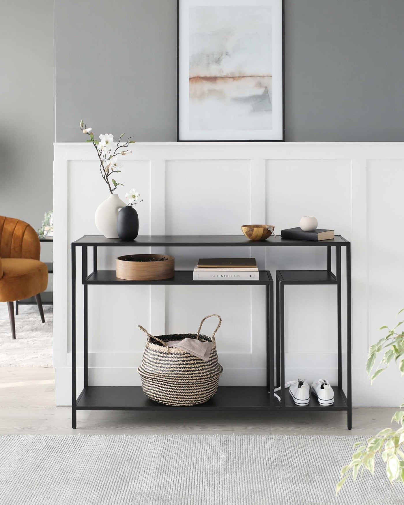 Modern two-tier console table with a matte black metal frame and sleek straight lines. The top tier displays a ceramic vase with white flowers, decorative bowls, and books. Below, a woven basket and white sneakers rest on the lower shelf. The table adds a functional yet stylish element to a contemporary interior with its minimalist design.