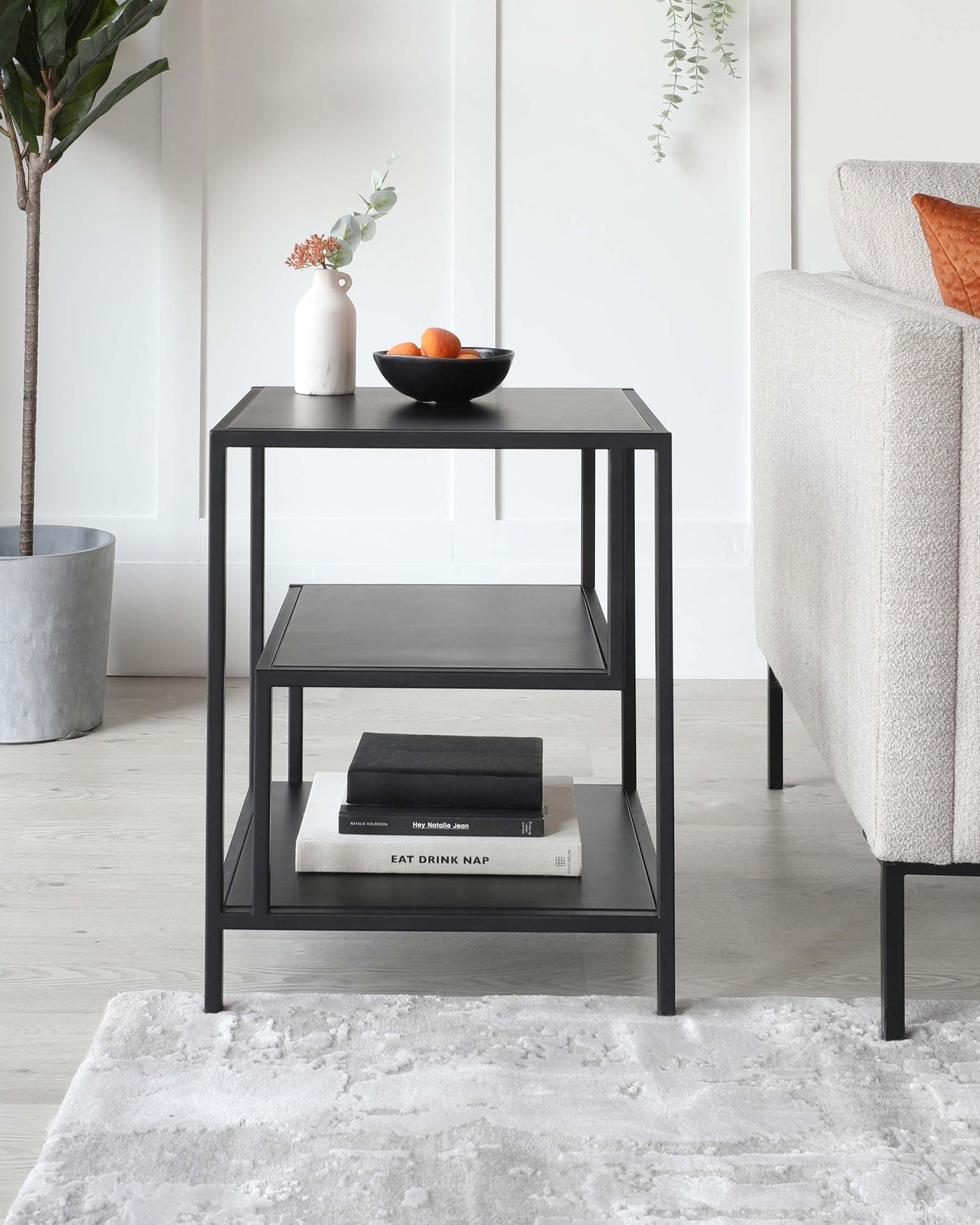 A modern, sleek three-tiered black side table with a minimalist design, displayed in a living area next to a sofa. The upper level holds a white vase with flowers and a black bowl with fruit, while the middle shelf contains neatly stacked books.