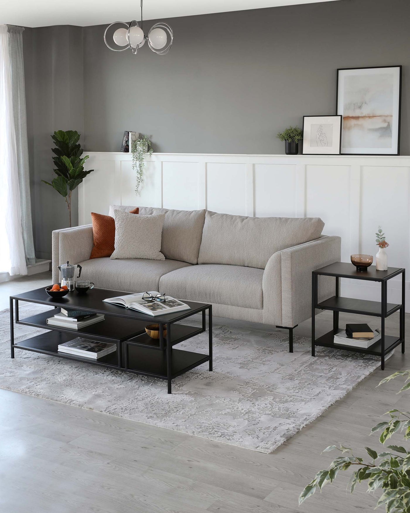 A contemporary living room setting featuring a textured fabric three-seater sofa in a light neutral colour, a large rectangular coffee table with a black metal frame and a dark wood finish top, complemented by dual lower shelves. Adjacent is a matching side table with a similar black metal frame and dual-tiered shelves, both featuring clean, modern lines. The furniture is arranged atop a soft, patterned area rug in muted tones, harmonizing with the room's modern yet cosy aesthetic.