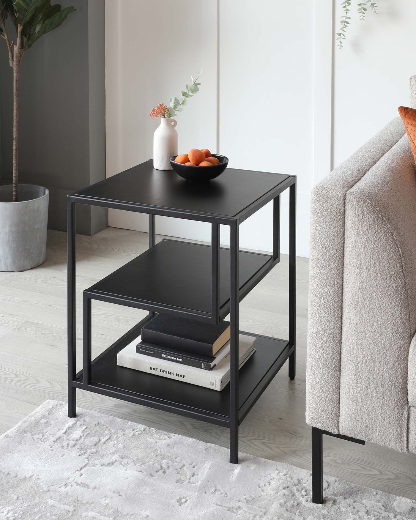 A modern, black, two-tiered square side table with a metal frame and a flat, square top surface, resting on four straight, square-cut legs. The lower shelf holds books, while the upper surface is styled with a white vase with flowers and a bowl of oranges. The table is set against a neutral background, with a grey upholstered sofa arm to the right, a houseplant to the left, and a light-grey area rug beneath.