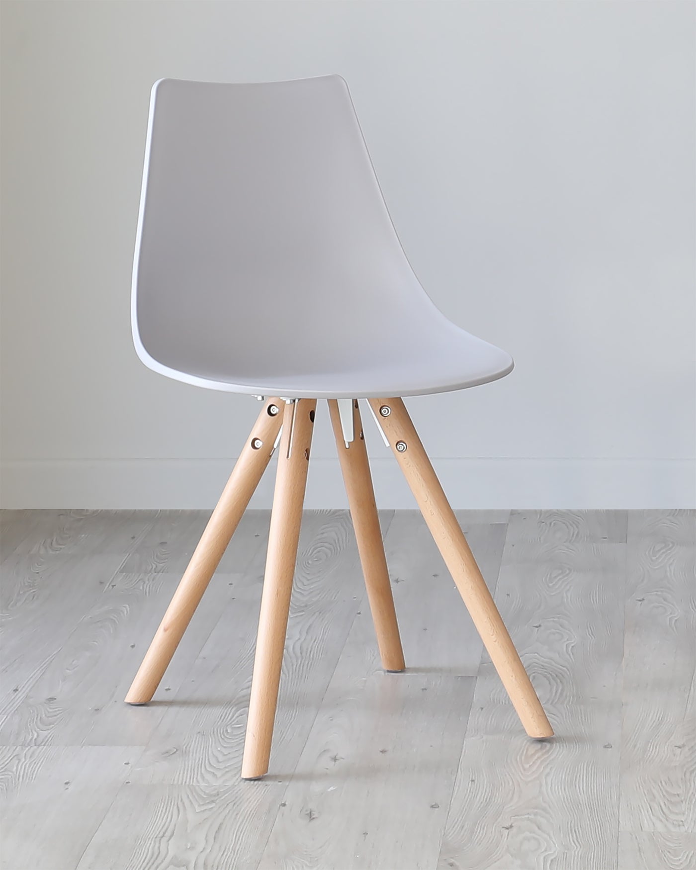 Modern minimalist chair with a white plastic seat shell and light wooden legs connected by metallic brackets, set against a plain wall on a light wooden floor.
