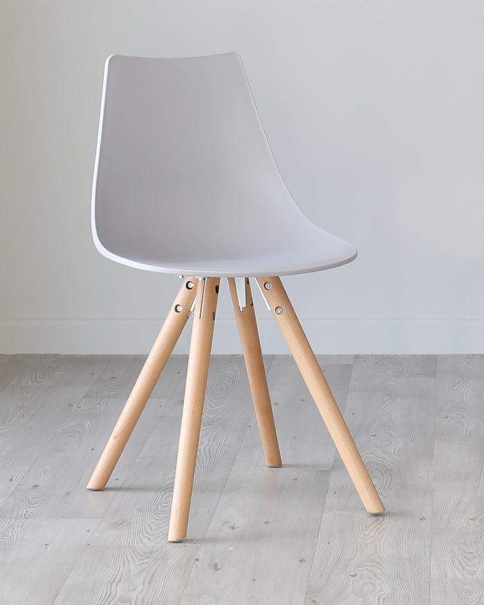 Modern minimalist chair with a white plastic seat shell and light wooden legs connected by metallic brackets, set against a plain wall on a light wooden floor.