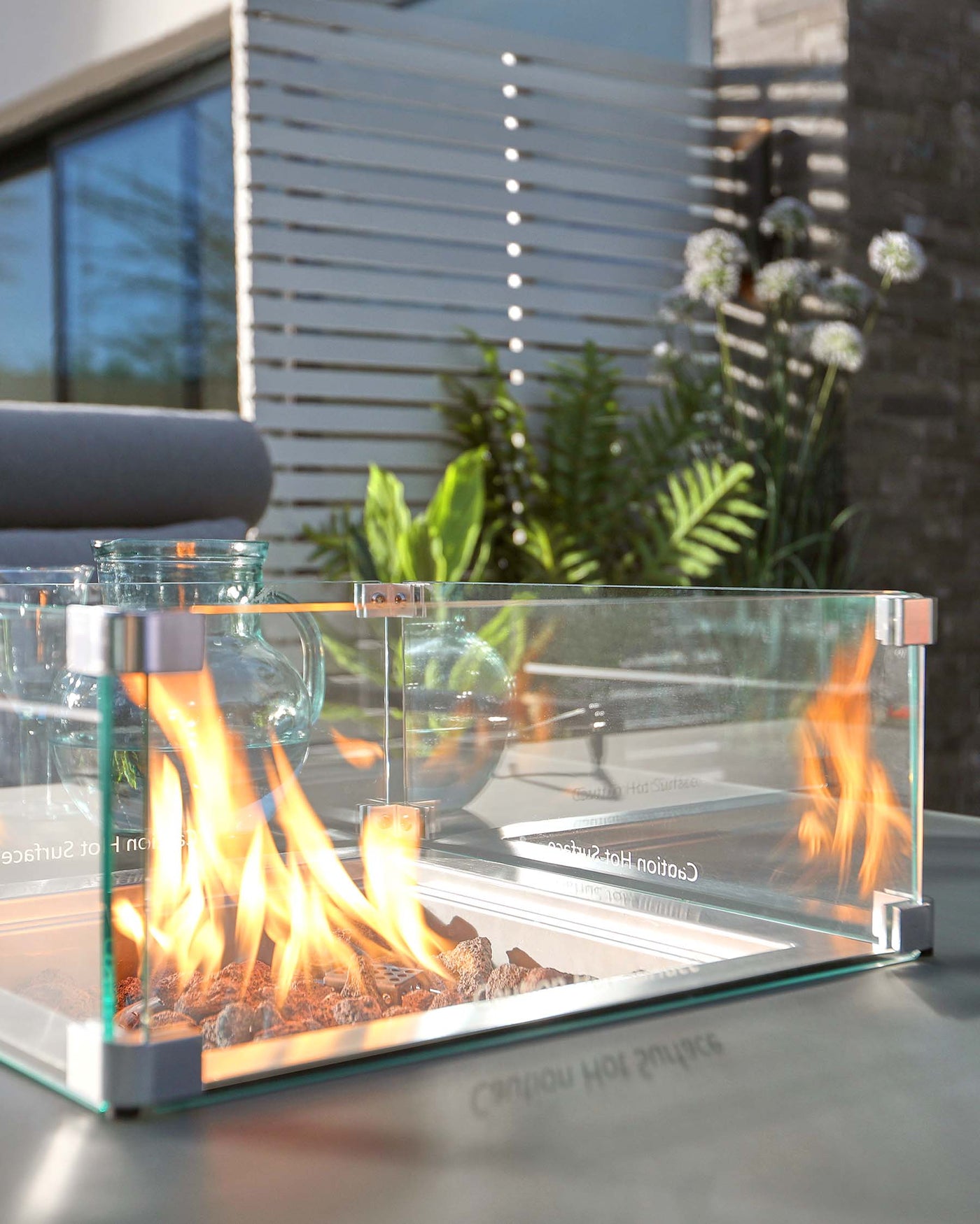 Modern outdoor glass fire pit table featuring a transparent fireproof glass barrier and polished metal corners, with flames burning over a bed of ornamental rocks. Visible in the blurred background is a sleek, contemporary patio setting with comfortable cushioned chairs and architectural features suggesting a high-end, minimalist design aesthetic.
