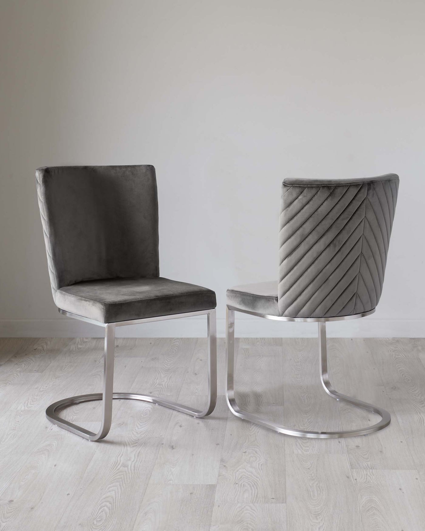 Two modern accent chairs with grey velvet upholstery and sleek silver metal cantilever bases. The chair on the right features an elegantly pleated backrest design, while the one on the left has a smooth finish. Both chairs display a minimalist style that merges comfort with contemporary aesthetics.