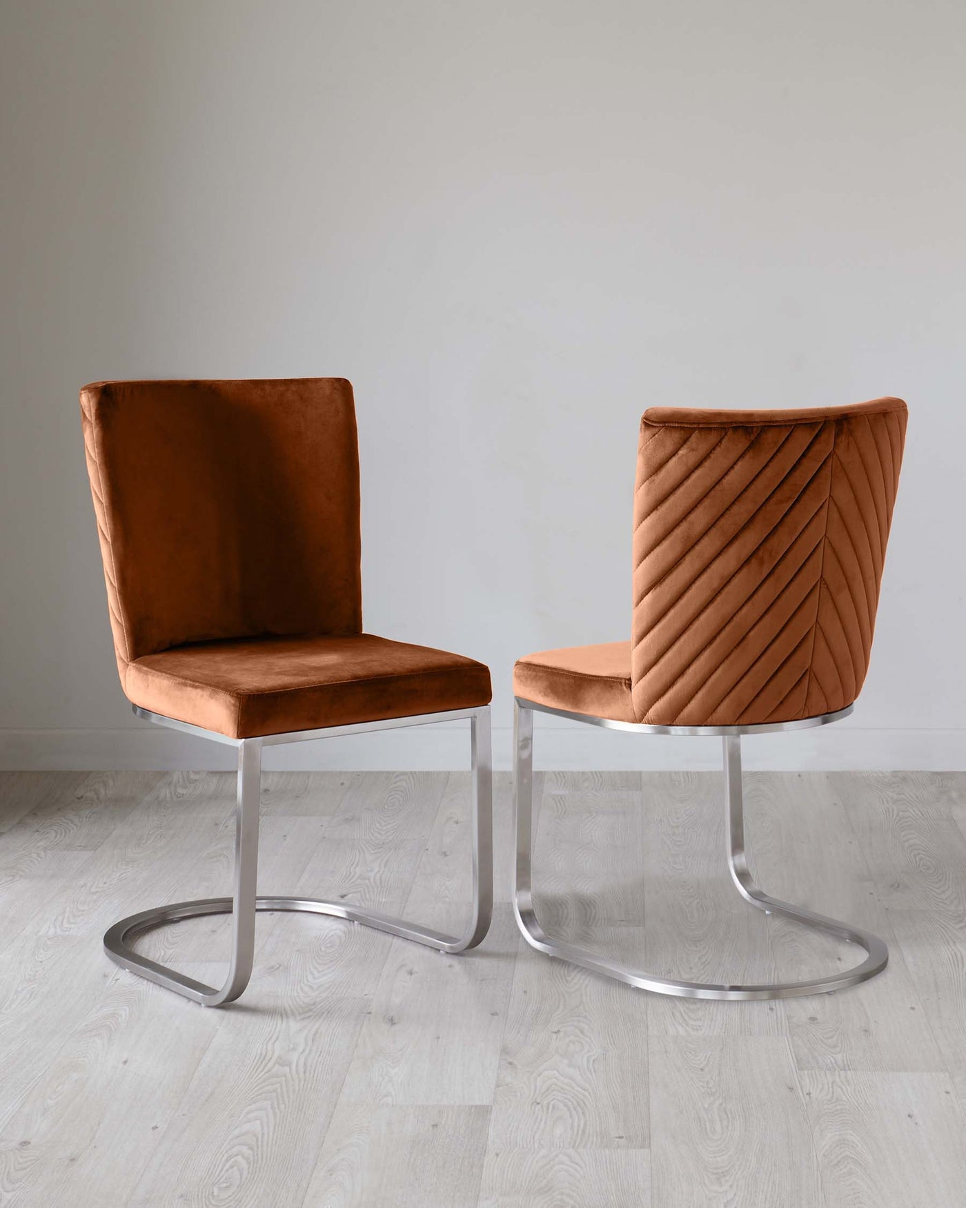 Two modern chairs with a sleek design, featuring burnished caramel velvet upholstery. The left chair has a clean, square back, while the right chair boasts a unique, diagonal pleat pattern on the backrest. Both have shiny, chrome cantilever bases, providing a contemporary touch on a light wooden floor against a neutral wall backdrop.