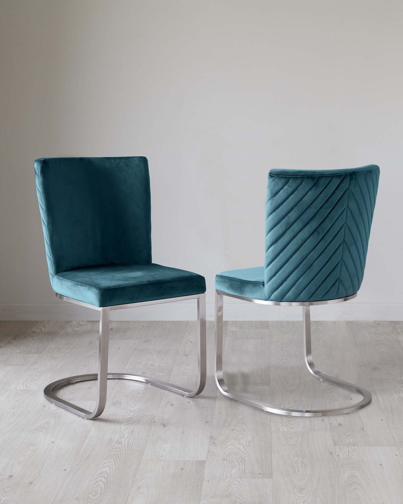 Two contemporary, teal velvet chairs with unique cantilevered metal bases against a neutral wall and light wood flooring. The chair on the right features an elegant pleated design on the backrest.