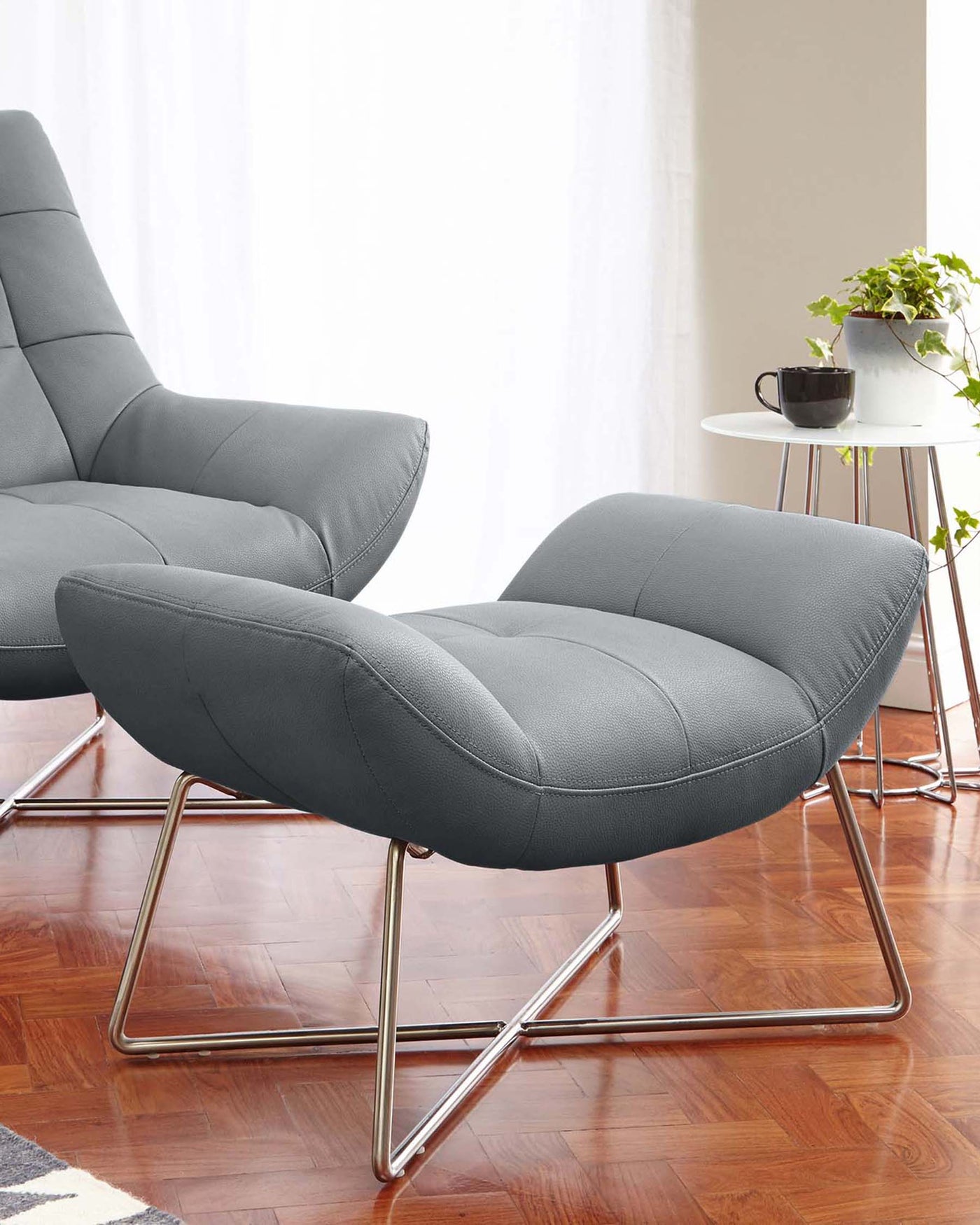 Modern grey upholstered lounge chair and matching ottoman with sleek chrome bases, positioned on a hardwood floor next to a small white round side table with a potted plant and mug.