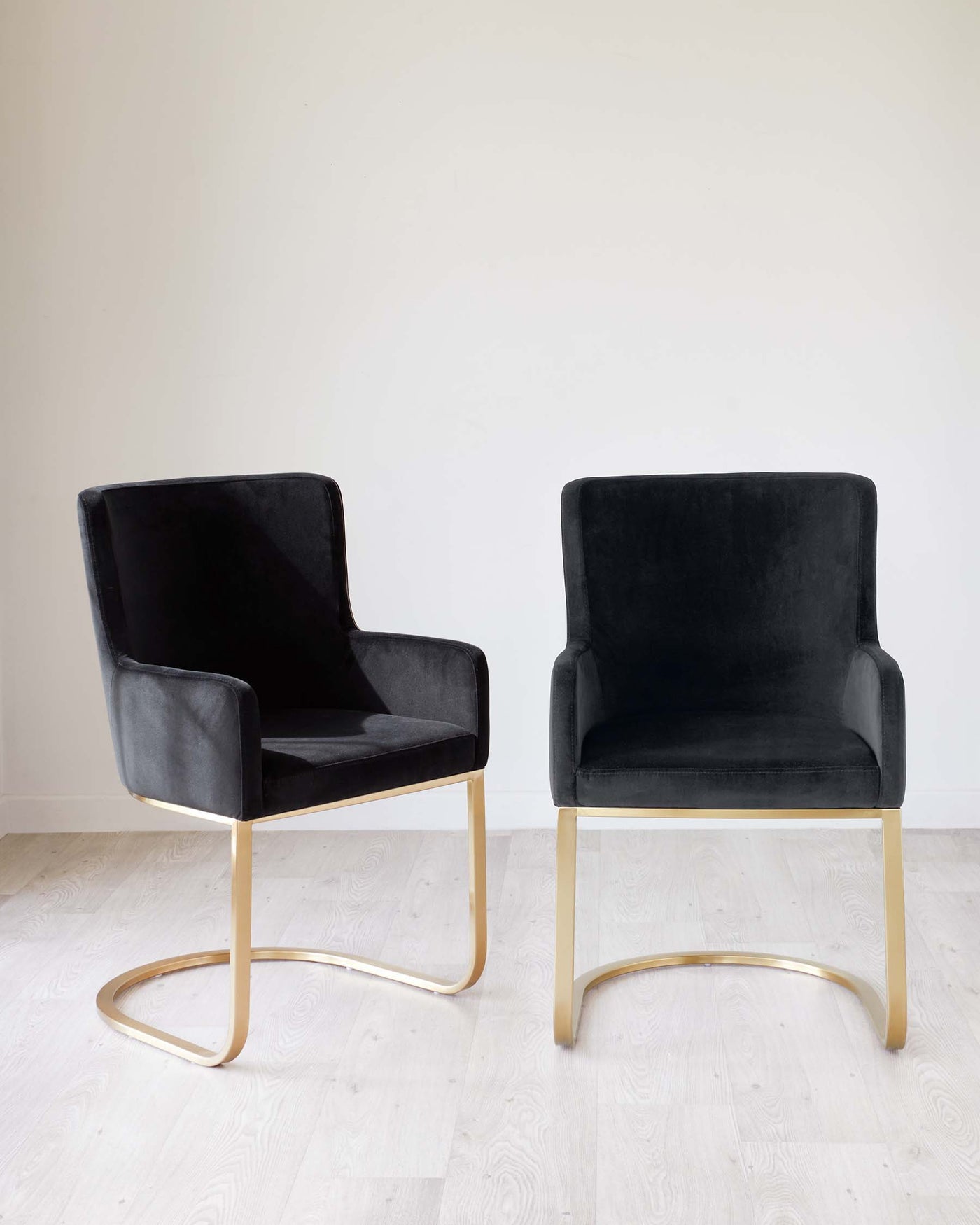 Two modern accent chairs with black velvet upholstery and sleek, gold-coloured metal legs, positioned against a white wall on a light wooden floor.