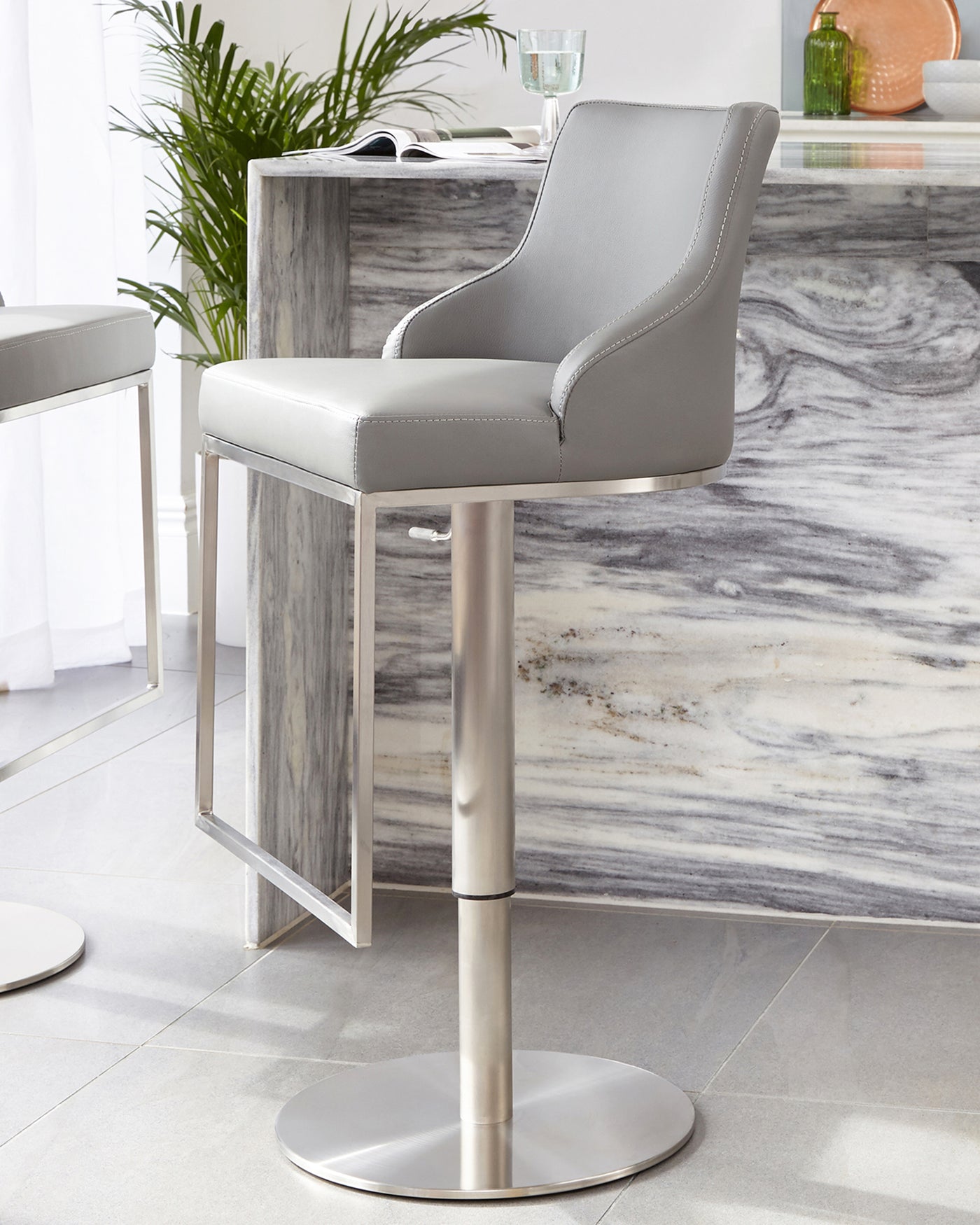 Modern grey upholstered barstool with a high back, winged sides, and silver nail head trim detailing. Features an adjustable-height chrome base with a built-in footrest and a wide circular foundation for stability.