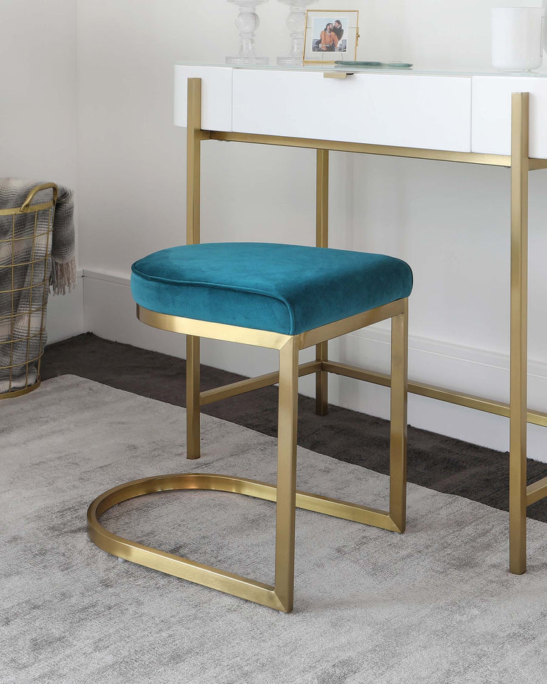 Elegant contemporary furniture set featuring a white console table with sleek gold-coloured metal legs and a matching velvet upholstered stool. The stool boasts a rich teal seat cushion and a distinctive oval-shaped gold metal base. The minimalist design is complemented by decorative items on the table, creating a chic and sophisticated ambiance.