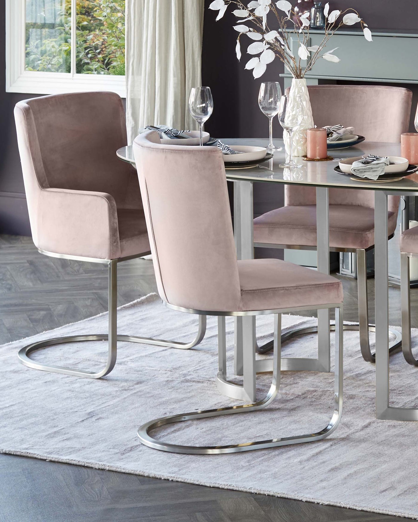 Elegant modern dining room furniture consisting of a clear glass-top table with sleek metallic silver legs and plush velvet-upholstered dining chairs in blush pink featuring unique curved chrome bases. The set is complemented by a soft grey area rug beneath.