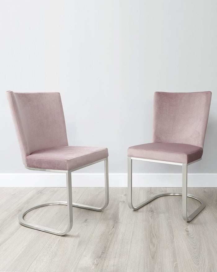 A pair of modern minimalist dining chairs with blush pink upholstery and sleek, cantilevered silver metal frames on a light wooden floor against a white wall.