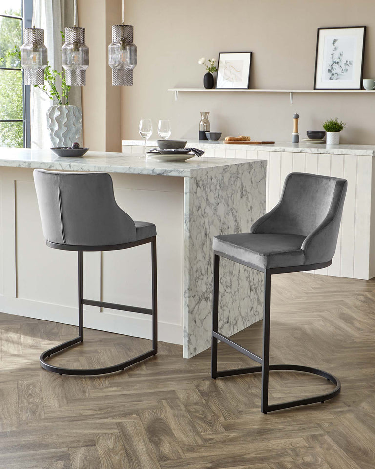 Two contemporary grey upholstered bar stools with a sleek metal frame, positioned at a marble-topped kitchen island with white shaker-style panelling.