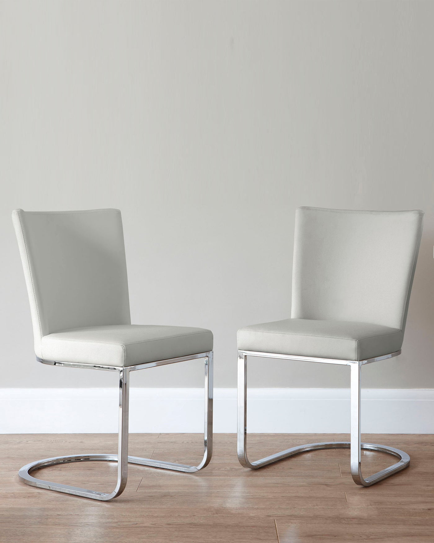 Two modern light grey upholstered chairs with sleek chrome cantilever bases, positioned on a wooden floor against a plain wall.