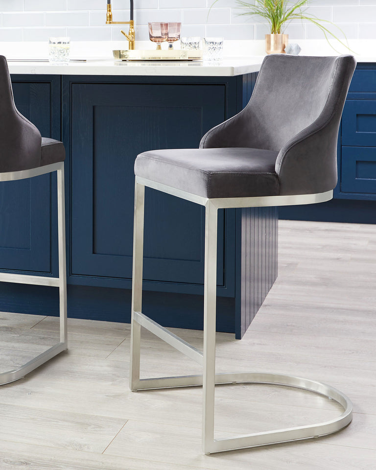 A pair of modern bar stools with grey velvet upholstery and a sleek silver metal frame, featuring a high back and a footrest for comfort.