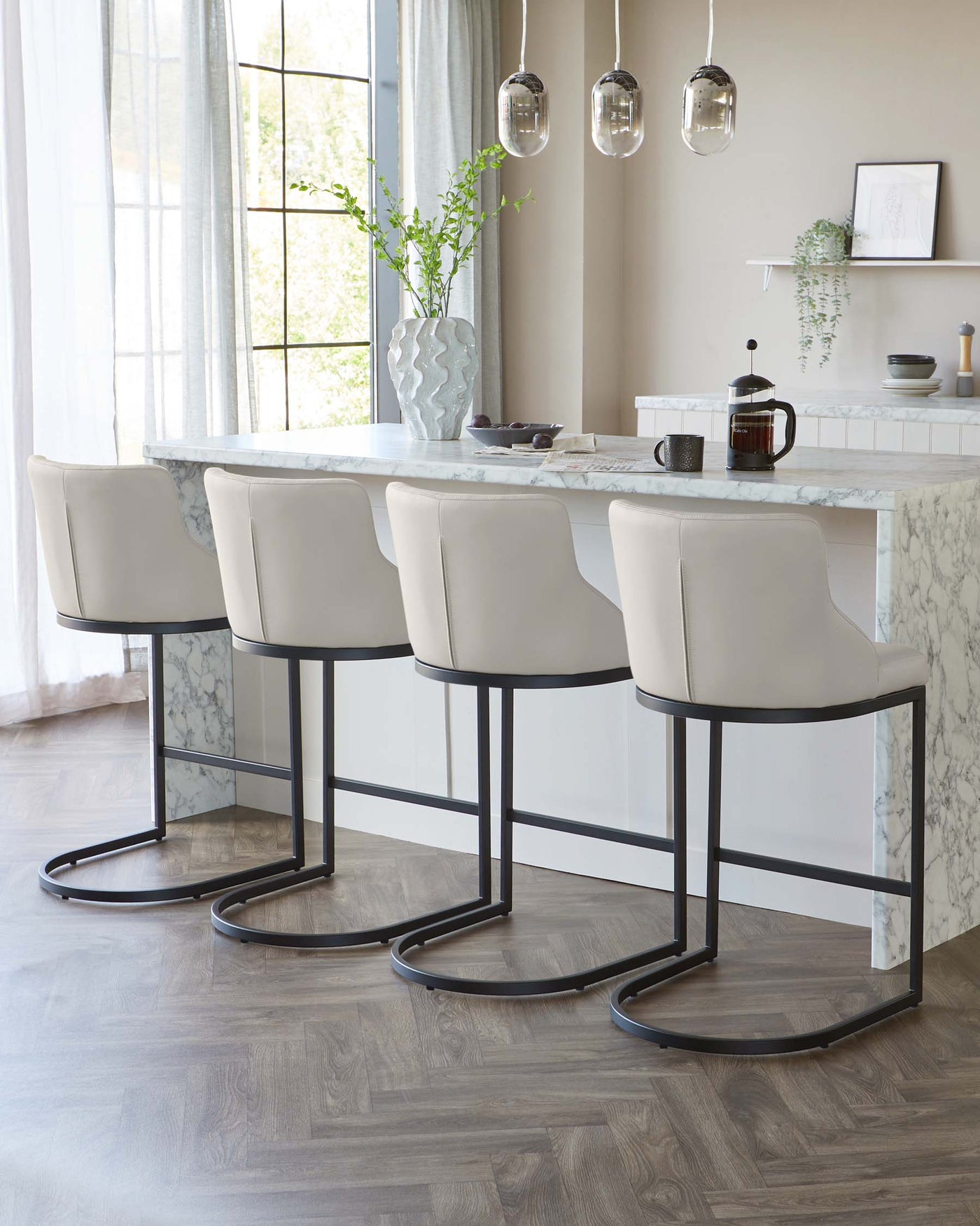 A set of four modern, elegant bar stools with cream-colored upholstered seats and curved backs, featuring slim black metal frames with a distinctive footrest design. Set along a marble kitchen island in a contemporary styled room.