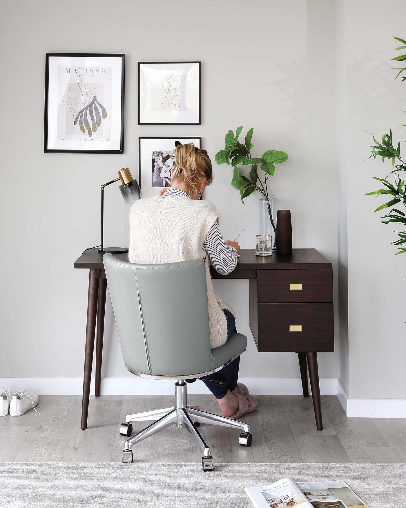 Modern home office setup featuring a sleek mid-century-inspired dark wood desk with tapered legs and brass-finished drawer pulls. Accompanied by a stylish grey upholstered swivel chair on a chrome star base with casters for mobility.