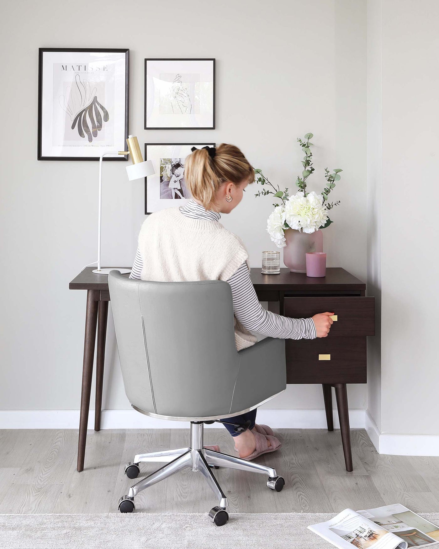 A contemporary dark brown wooden desk with tapered legs and two drawers paired with a modern grey office chair on casters with a high back and chrome base.