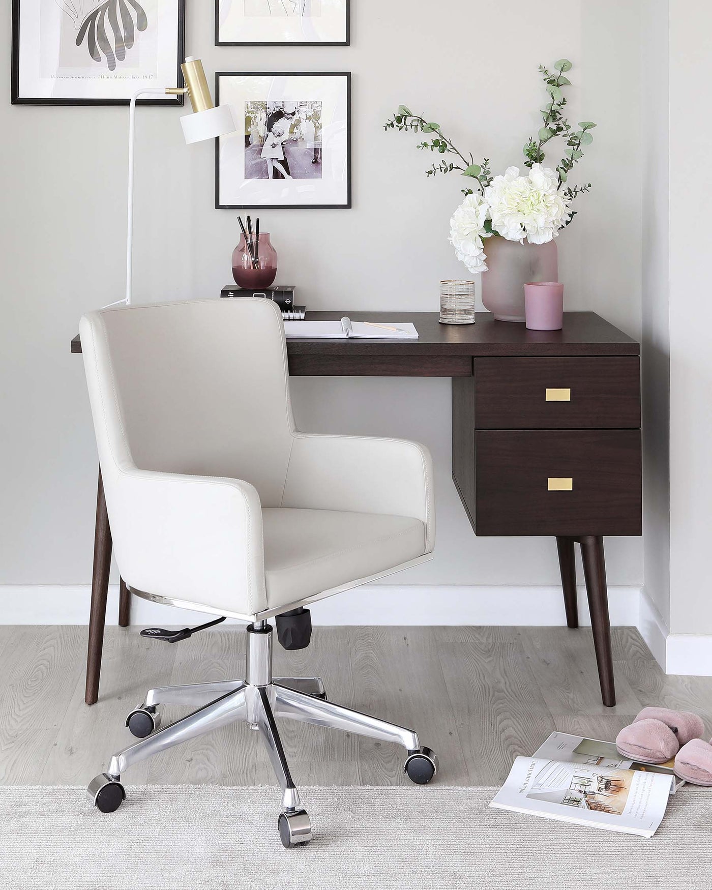 Modern minimalist home office setup featuring a sleek dark wood desk with a slim drawer and angled legs, paired with an elegant white leather office chair with a high back and chrome swivel base. The desk is accessorized with a brass desk lamp, framed artwork above, and a vase with white flowers for a touch of organic warmth. A light-coloured rug under the chair softens the look of the light wood flooring.