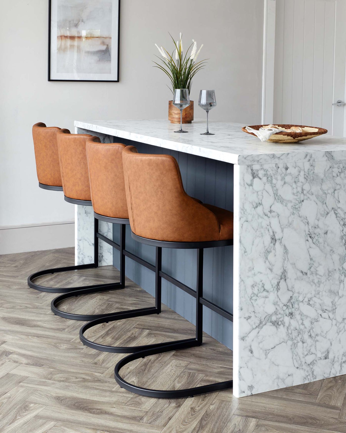 Contemporary kitchen bar area featuring a sleek white marble countertop with overhang for seating, complemented by three stylish caramel brown leather high-back barstools with black metal frame legs set on herringbone pattern wooden flooring.