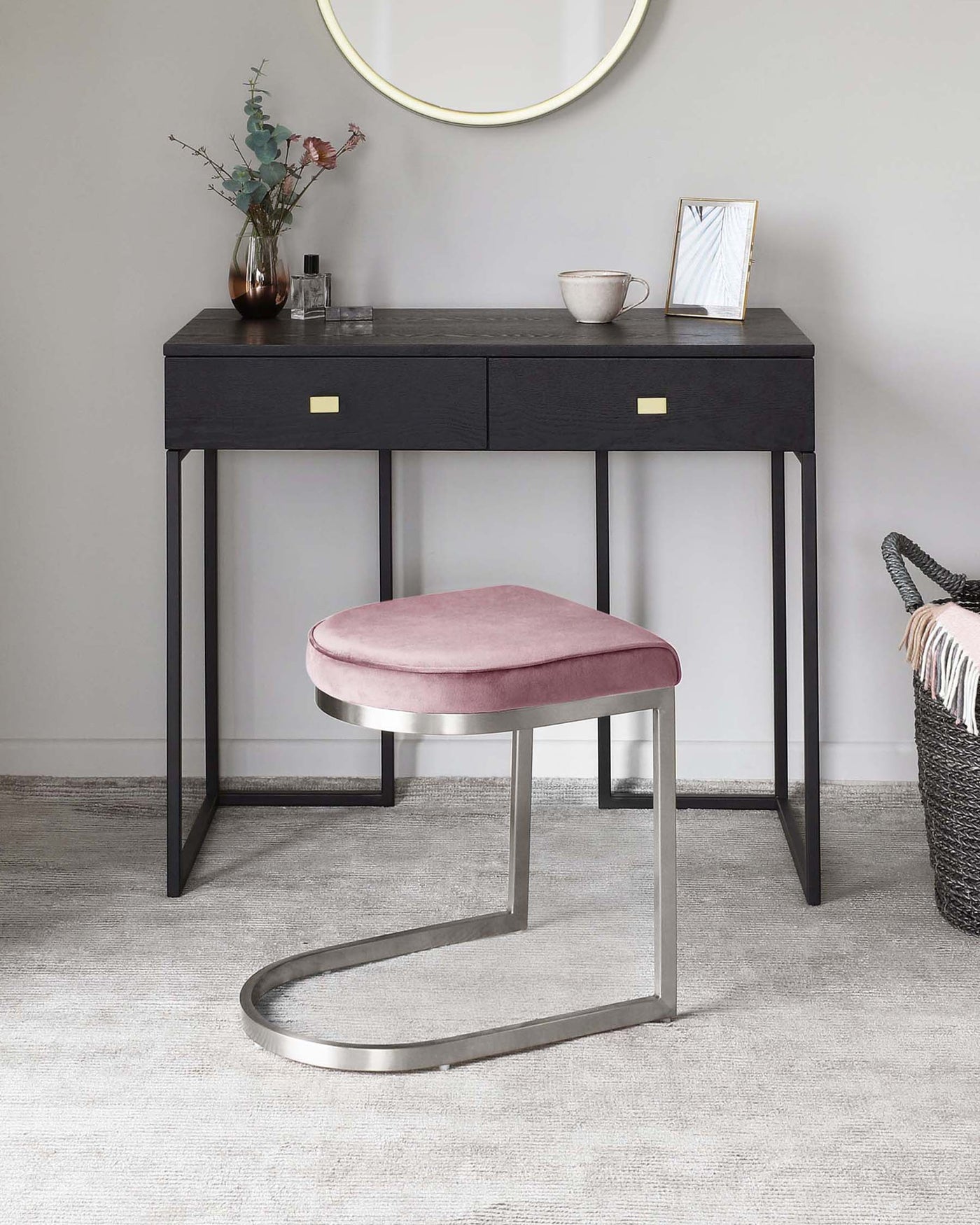 Black wood console table with slender metal legs and two drawers featuring brass handles, paired with a round mirror above and a modern stool with a velvety pink cushion and silver metal base.