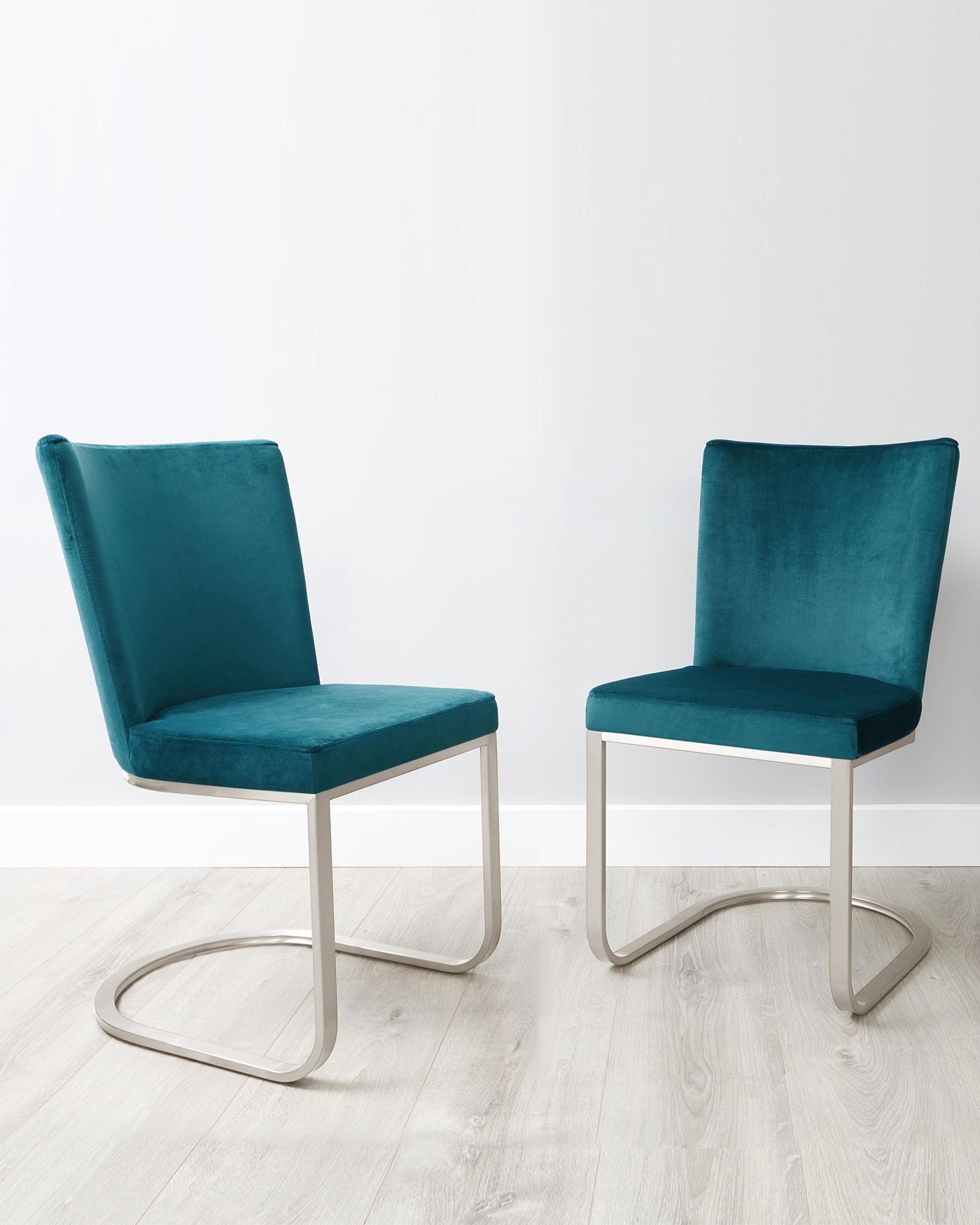 Two contemporary teal velvet dining chairs with sleek silver metal cantilever bases, on a light wooden floor against a white background.