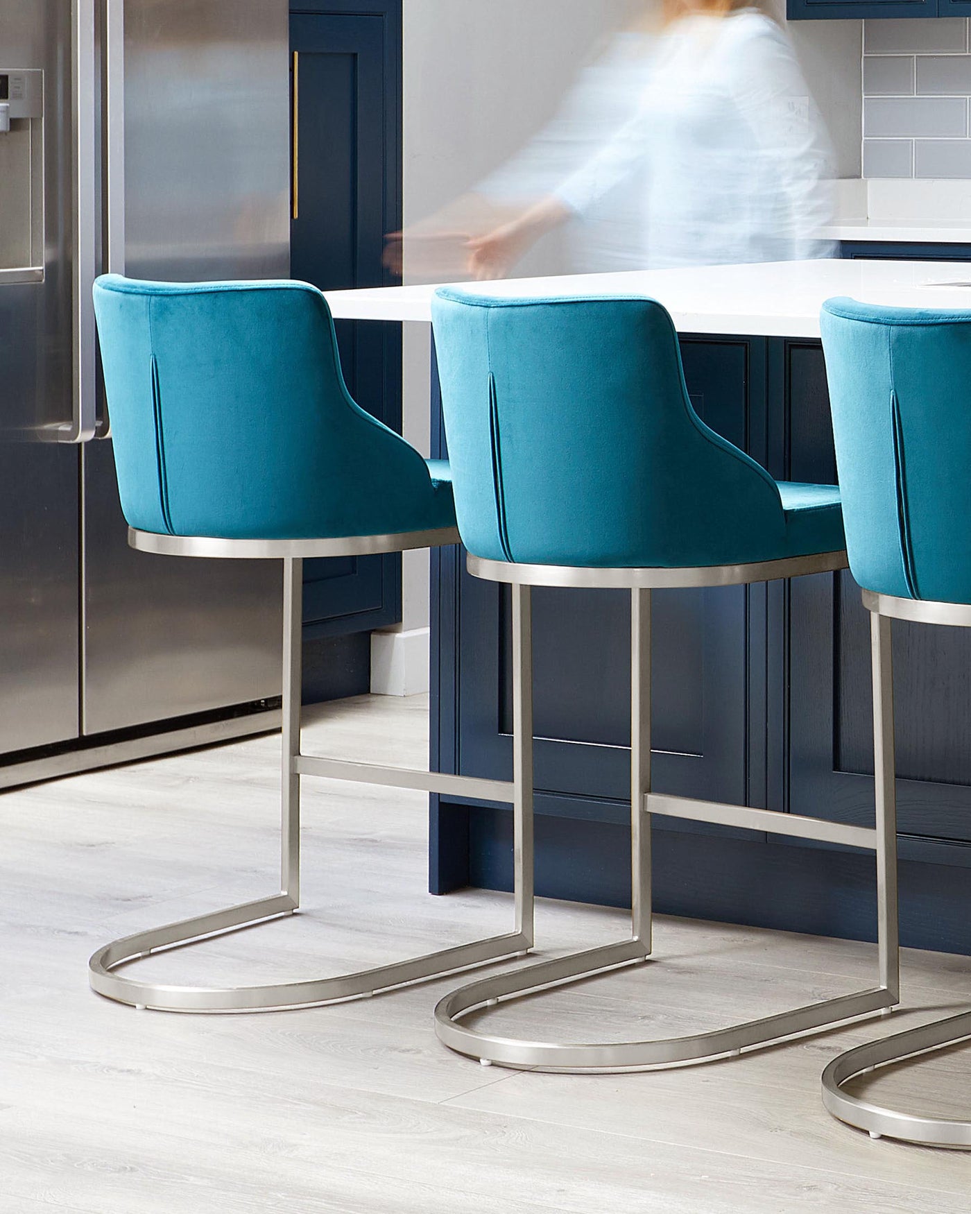 Teal blue upholstered bar chairs with a sleek curved back design and vertical stitching detail. The chairs feature a silver metal base with flat rounded legs and a circular footrest. The furniture is set against a kitchen island with a white countertop.