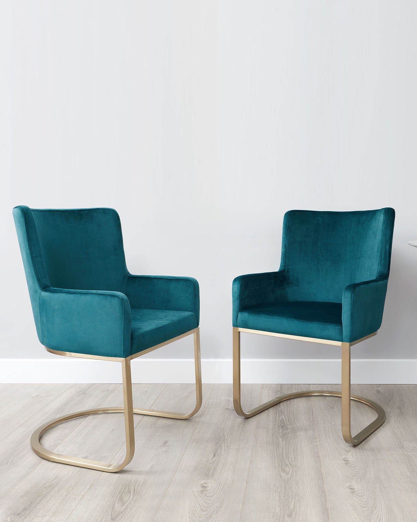 Two modern accent chairs with plush teal velvet upholstery and sleek gold-finished metal legs, set on a light hardwood floor against a white wall.