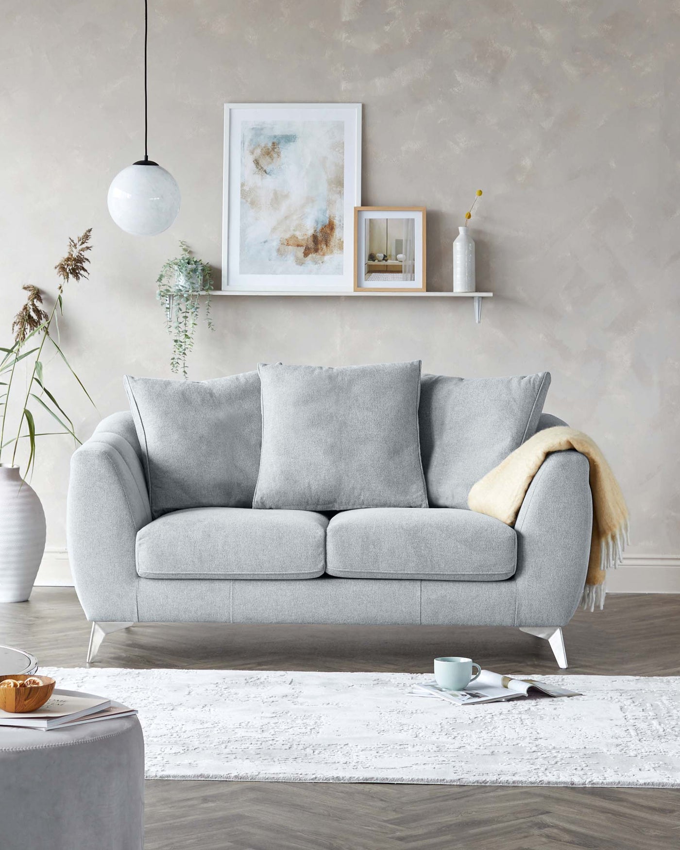 Elegant modern two-seater sofa with plush grey fabric upholstery, featuring deep cushioned seats, a gently curved backrest, and sleek, minimalistic metal legs. A soft beige throw is draped on one side, adding a touch of warmth to the otherwise cool tones of the room. The sofa is complemented by a textured white area rug beneath it, creating a cosy and stylish living space.