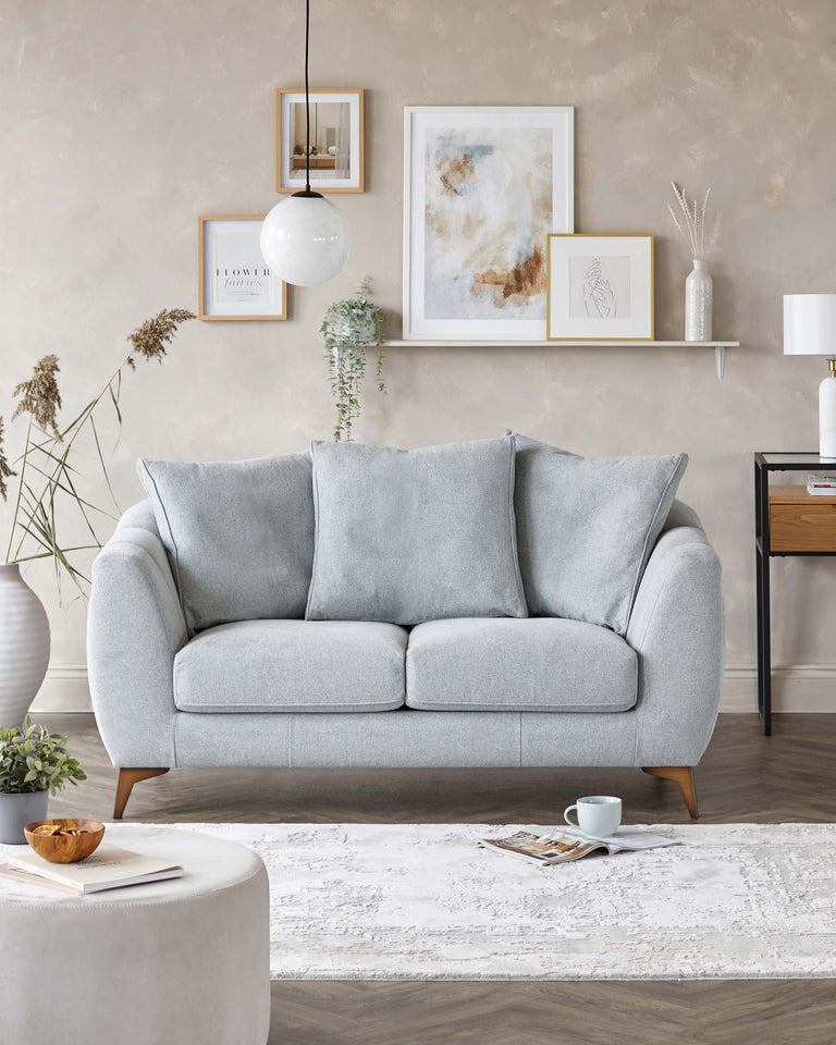 A modern light grey fabric three-seater sofa with cosy cushions, tapered wooden legs, and a slightly flared armrest design. In front of the sofa is an off-white round ottoman on a white textured area rug. To the side is a black end table with open shelving and a table lamp with a white shade. The background includes decorative wall art and a hanging globe pendant light.
