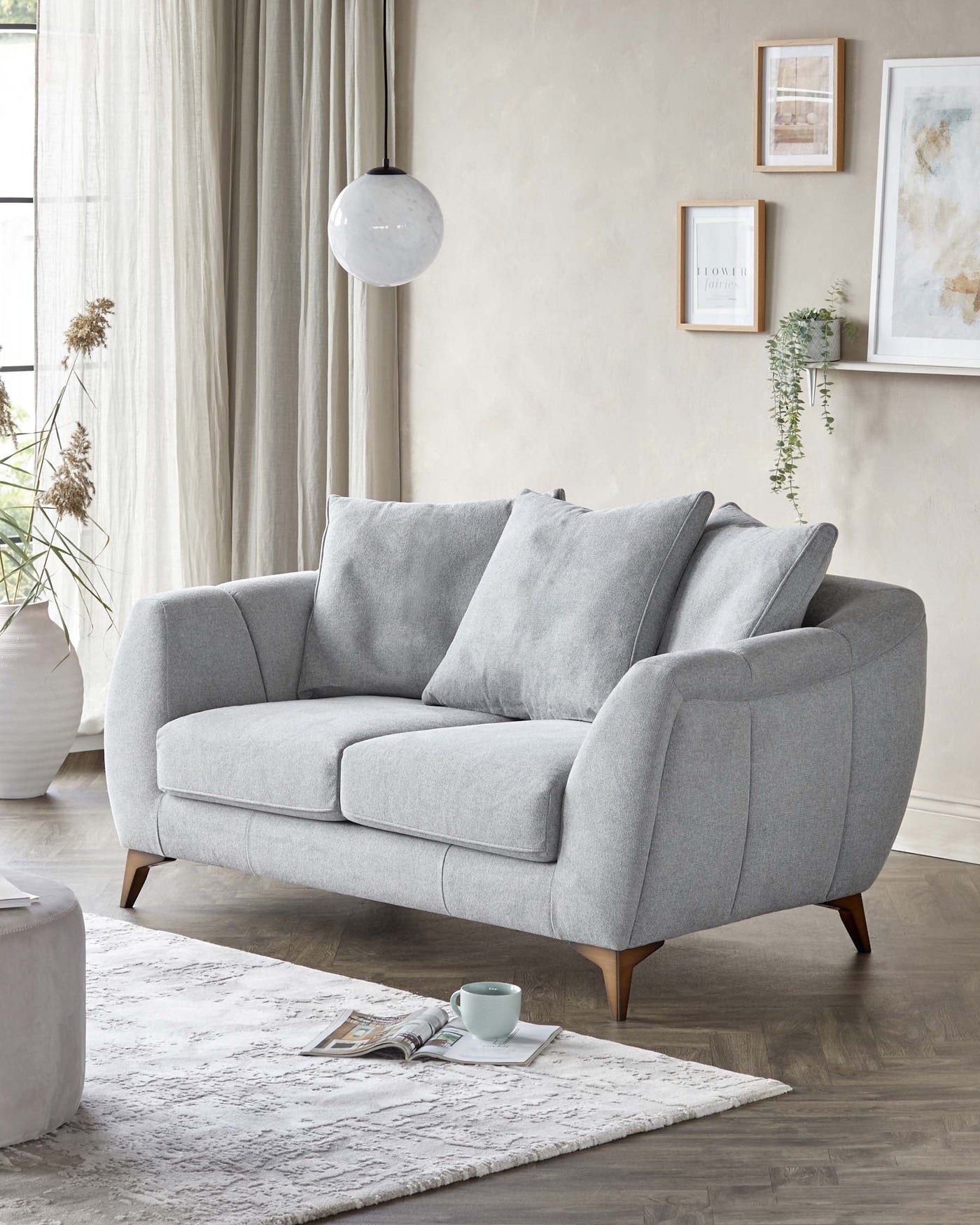 A modern mid-century-inspired grey fabric sofa with a curled armrest design, featuring plush back cushions and a tufted seat cushion, elevated on tapered wooden legs. In front of the sofa, there's a round, light grey ottoman, and underneath is an off-white textured area rug.