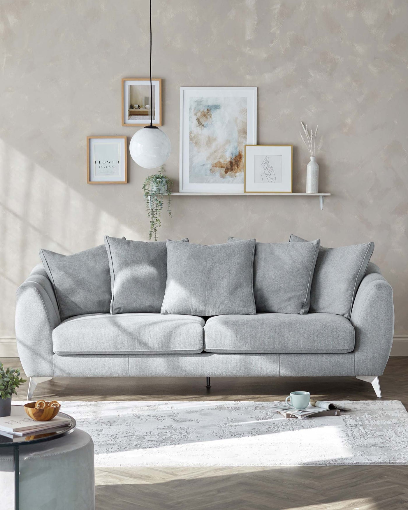 A modern light grey fabric sofa with plush back cushions and a tufted seat cushion resting on low-profile metal legs. The sofa is adorned with matching grey throw pillows and is set against a textured beige wall with decorative floating shelves and framed artwork above. A minimalist white area rug lies beneath the sofa, accompanied by a sleek round side table and a casually placed magazine and tea set on the floor.