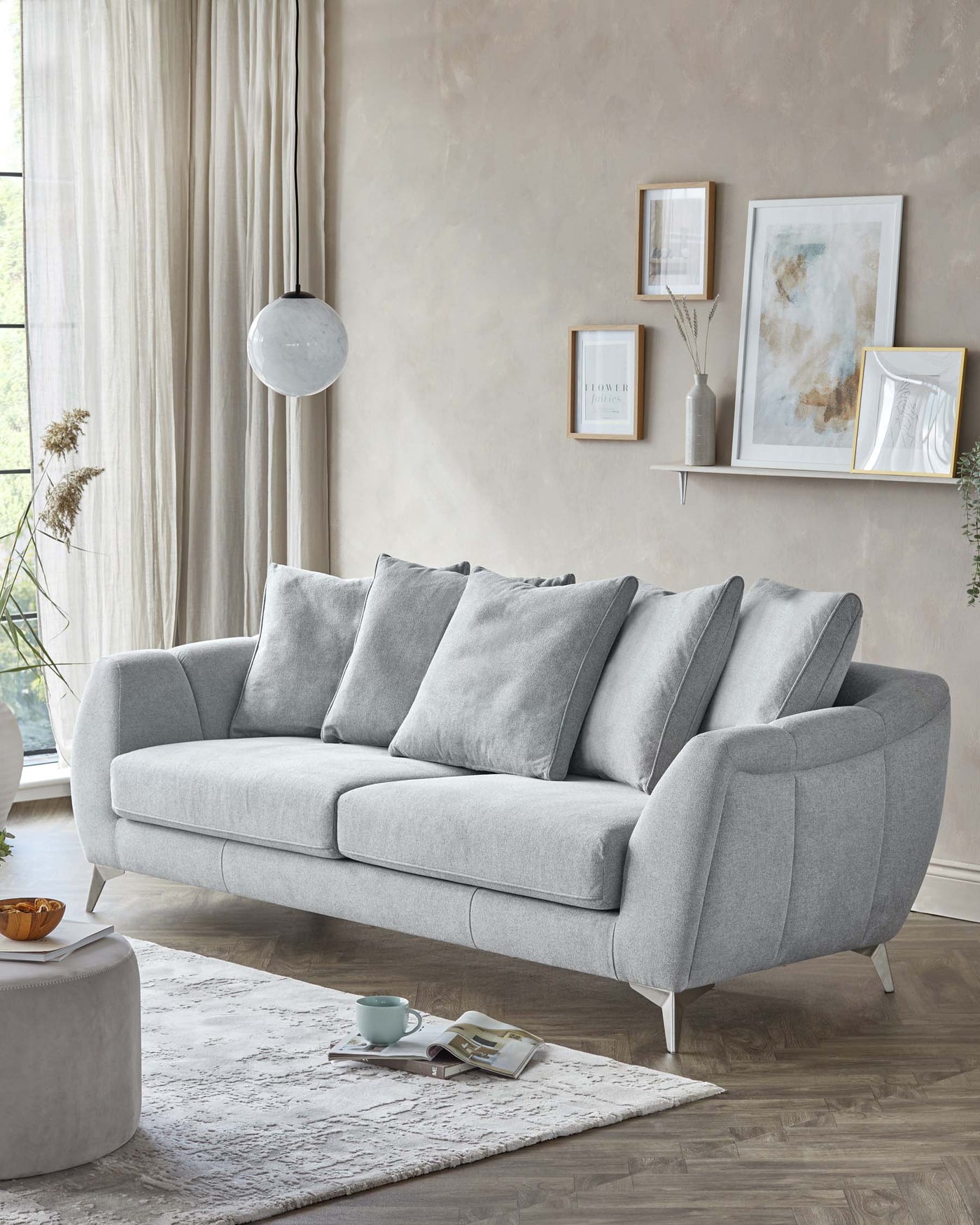 Elegant modern living room setup featuring a light grey three-seater sofa with plush back cushions and sloping armrests, complemented by angled wooden legs. A circular, light grey fabric ottoman sits on an area rug in front of the couch, alongside a small stack of books and a ceramic tea cup on a saucer.