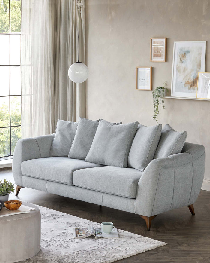 A modern mid-century inspired sofa with a gently curved silhouette, featuring light grey upholstery, cylindrical cushions, and angled wooden legs. A cylindrical light grey fabric ottoman with nail head trim accents is positioned on a textured off-white area rug in front of the sofa.