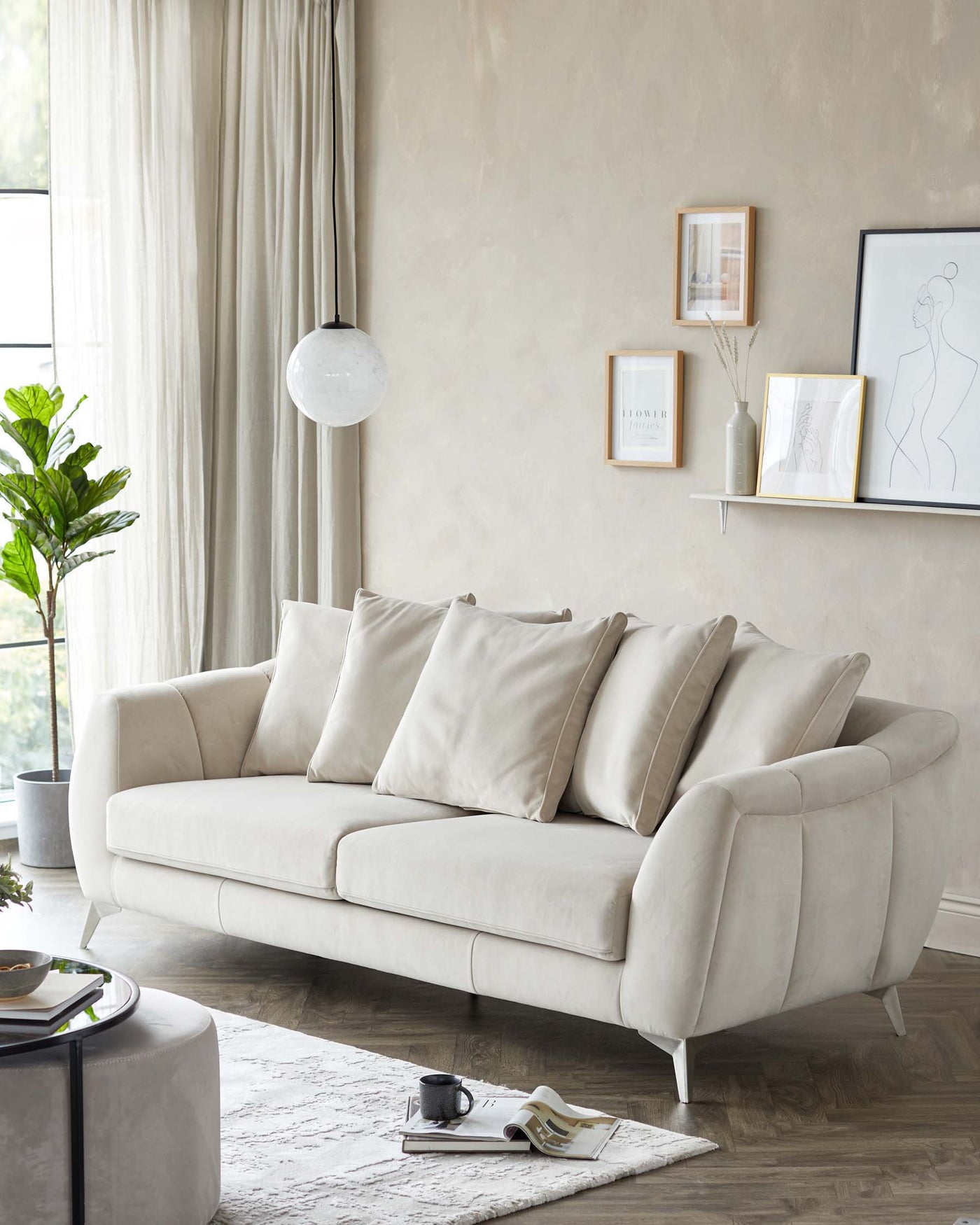 Elegant contemporary three-seater sofa with plush beige upholstery, featuring a curved arm design, accompanied by matching beige scatter cushions. A chic round grey ottoman sits as a centrepiece on a textured white area rug, with reading materials casually placed on it. The setting is complemented by minimalist decor and natural lighting, creating a serene living space ambiance.