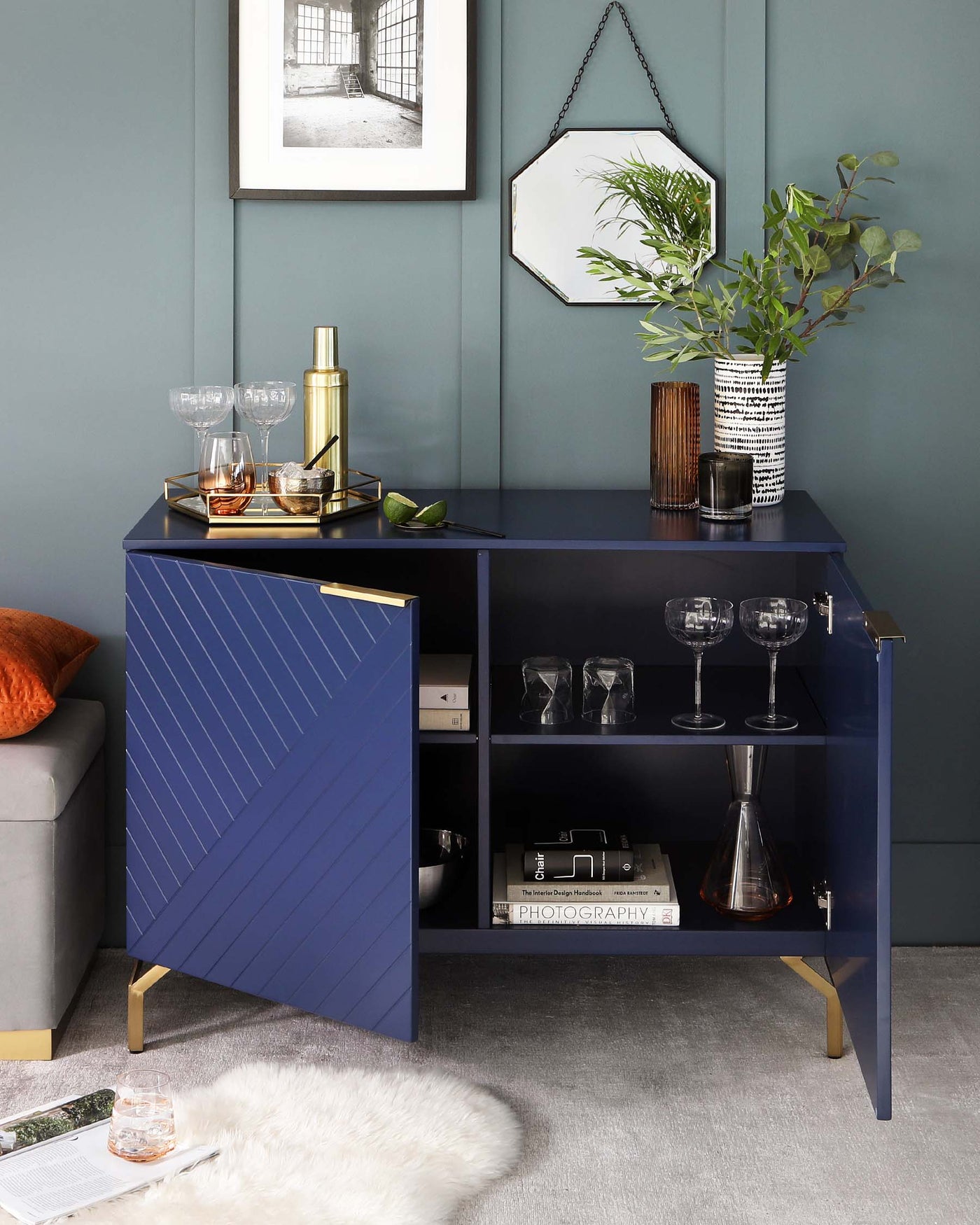 Modern navy blue bar cabinet with gold accents and chevron pattern on the front door, featuring an open shelf with books and glassware, and a closed compartment. Elegant metal legs support the structure. The cabinet is accessorized with a cocktail mixing set on a tray, a decorative vase with greenery, and a framed picture and hexagonal mirror adorn the wall above it.