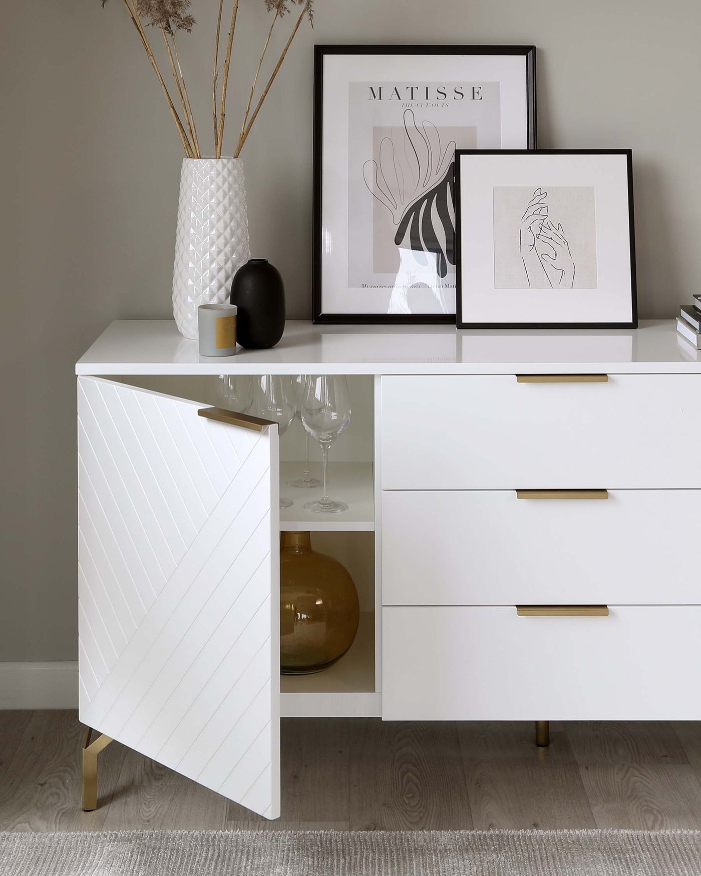 Modern white sideboard with chevron-patterned door detailing, complemented by sleek brass handles and matching brass feet, featuring two visible drawers and partially visible shelving space.