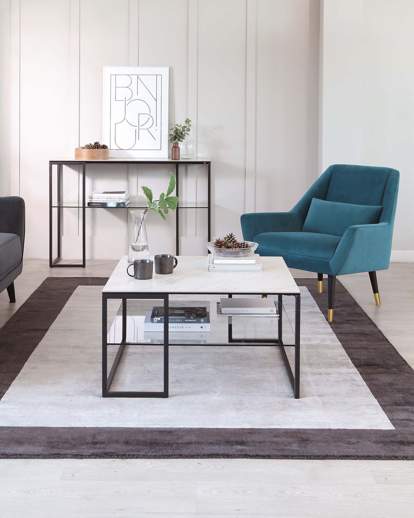 Modern furniture set in a minimalist living room including a teal blue armchair with tapered legs and soft fabric upholstery, a sleek rectangular coffee table with a white top and black metal frame, and a slim console table with a matching black metal frame and wooden top. The room features neutral tones, with the furniture placed on a gradient grey area rug. Decorations include framed wall art, a carafe and glasses on the coffee table, and plant accents on the console table.
