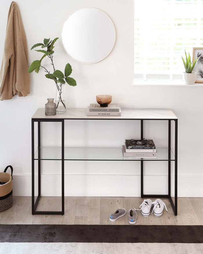 Minimalist black metal frame console table with a rectangular stone top and a lower glass shelf, positioned against a white wall with decorative items and adjacent to a round mirror.