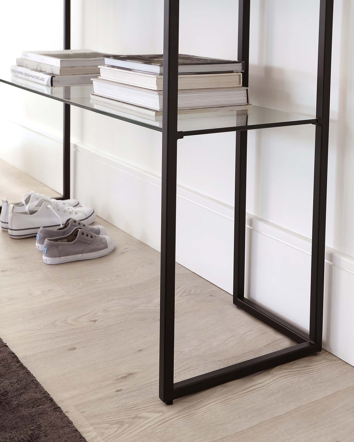 A minimalist black metal frame shelving unit with three levels, featuring clear glass shelves filled with neatly stacked books. The unit is positioned against a light-coloured wall above a light wooden floor, with a pair of sneakers casually placed to one side.